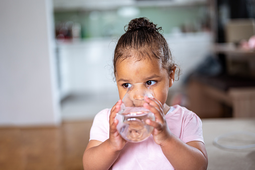 Little cute african american girl drinking water from the glass in a domestic room.