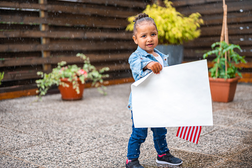 Two years old cute little african-american girl, wearing white t-shirt and jeans jacket, standing in the rain and holding an american flag and a white poster. Wooden fence and flower pots in the blurred background.