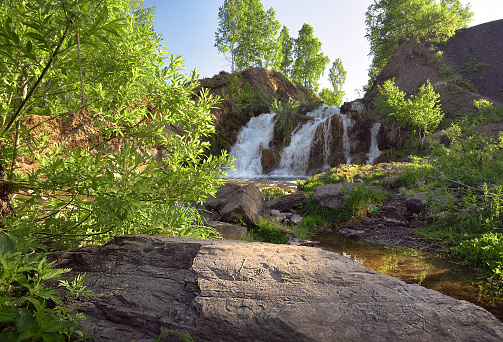 A small waterfall among the rocks, surrounded by birch trees with spring foliage in the rays of the sun. White streams of water, desolation, fresh greenery, large stones, the nature of Siberia. Novosibirsk region, Russia, 2020