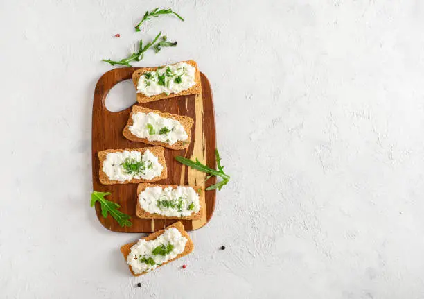 Crispy bread crackers with cottage cheese and green herb on wooden cutting board on white background. Top view. Copy space.