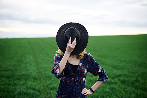 Young blond woman, wearing long dark boho dress, holding black hat covering face on green field in spring. Model posing outside in meadow. Hippie musician at natural environment.