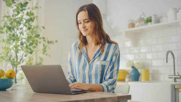 istock Young Beautiful Woman Using Laptop Computer While Wearing Blue Pyjamas. Brunette Female Sitting in a Modern Kitchen Room. Working from Home During Quarantine. 1248941686