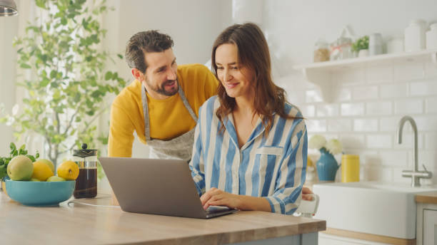 istock Beautiful Young Couple Using Laptop Computer in Kitchen at Home. Female Wearing Pyjamas. Man Wearing an Apron is Encouraging Girlfriend with a Shopping Choice. Female Sitting and Browsing Internet. 1248941704