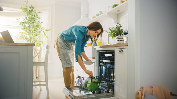 beautiful female está carregando pratos sujos em uma máquina de lavar louça em uma cozinha ensolarada brilhante. garota usando um avental. jovem dona de casa usa aparelho moderno para manter a casa limpa. - contemporary indoors lifestyles domestic room - fotografias e filmes do acervo