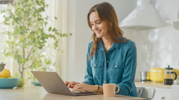 istock Young Attractive Woman Using Laptop Computer and Drinking Morning Cup of Coffee While Wearing a Jeans Shirt. Brunette Female Sitting in a Modern Cozy Kitchen. Freelancer Working from Home. 1248942859