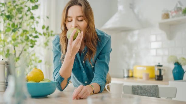 autentico ritratto di bellezza di una bella femmina bruna che mangia una grande mela verde in una stanza luminosa a casa. pretty young woman posa per la fotocamera e sorride delicatamente. vegetariano naturale sano. - apple women green eating foto e immagini stock