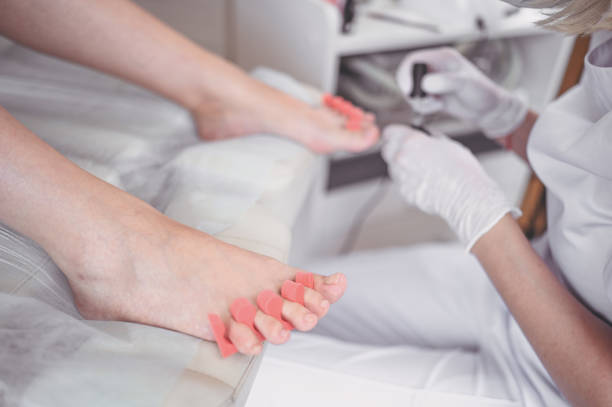 istock Pedicurist doing white nail polish on client legs using toe finger separator. Professional medical pedicure procedure. Foot treatment in SPA salon. Podiatry clinic. Beautician hands in white gloves. 1248943348