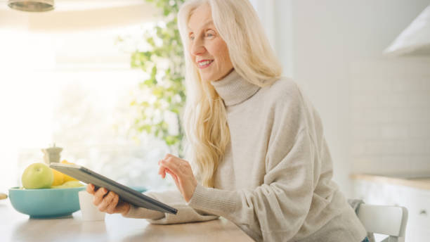 istock Authentic Senior Woman Using Tablet Computer in Kitchen Room at Home. Beautiful Old Female Pensioner with Gray Hair Writes a Text in a Messenger on a Device. Elderly Person in Hand with Technology. 1248945035