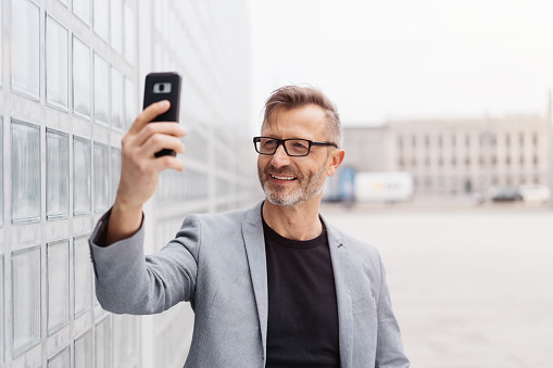 Street portrait of cheerful mature man wearing grey jacket taking selfie