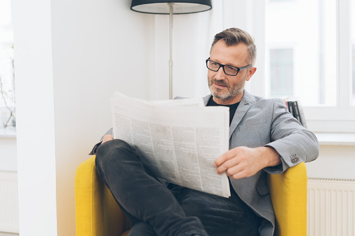 Portrait of mature man wearing glasses reading newspaper while sitting in yellow armchair