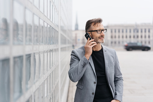 Businessman wearing glasses standing alongside a modern commercial building talking on a mobile phone in town looking off to the side