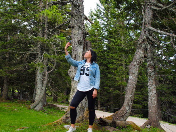 Woman Taking Selfies in the Woods Woman with black hair , wearing jeans jacket, taking selfies in forest, green pine trees in tha back krvavec stock pictures, royalty-free photos & images