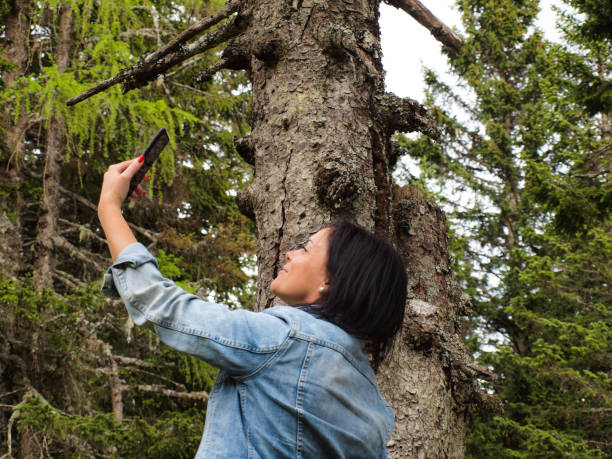 Woman Taking Selfies in the Woods Woman with black hair , wearing jeans jacket, taking selfies in forest, green pine trees in tha back krvavec stock pictures, royalty-free photos & images