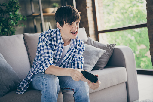 Photo of teenager school boy sit comfy couch stay home quarantine time hold joystick, playing video game addicted gamer excited mood trying hard living room indoors