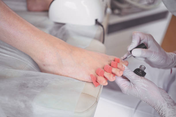 pedicurist che fa smalto bianco sulle gambe del cliente usando lampada a conchiglia. procedura di pedicure medica professionale. trattamento del piede nel salone spa. clinica di pododiatria. il medico estetista consegna i guanti bianchi. - pedicure podiatrist human foot moisturizer foto e immagini stock