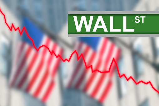 Wall Street sign post with red line chart diagram indicating stock market going down. In the background blurred American US flags hanging from buildings in New York City.
