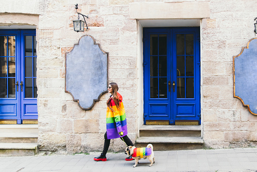 Woman with long hair in sunglasses and her small cute dog - pug breed in colorful raincoats walking in the old city enjoying the bright sunny spring day