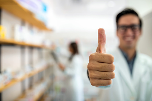 Friendly smiling male pharmacist in white overall posing with thumbs up in drugstore. Success in the medical world
