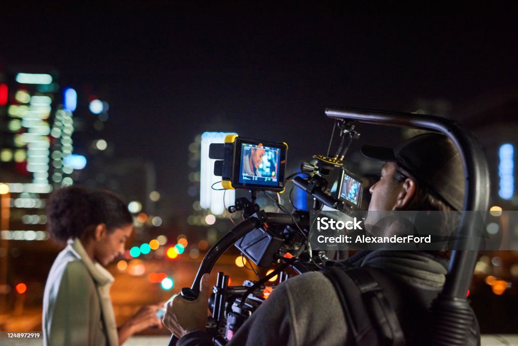Camera, lights, action! Behind the scenes shot of a camera operator shooting a scene with a businesswoman at night Filming Stock Photo