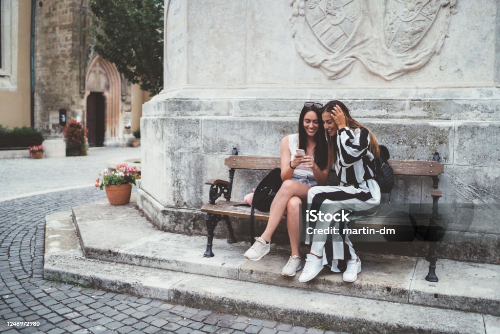 Friends traveling around Europe Tourists in Budapest sitting at bench, surfing the net on smartphone Sopron Stock Photo