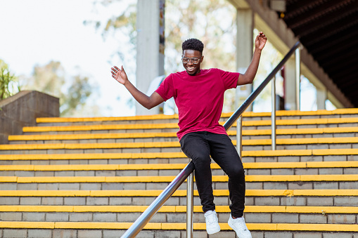 A black male student in his 20's sliding down the railing of some steps with his arms wide open and a smile on his face.