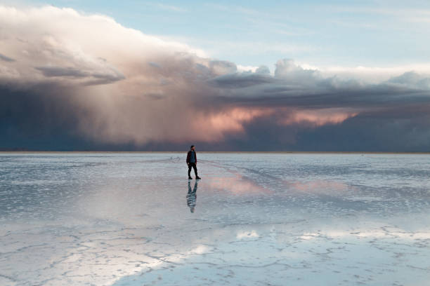 Tourist in red checkered shirt is walking in Salar de Uyuni at the sunset with dramatic sky and mirror reflection in the water. Epic view.