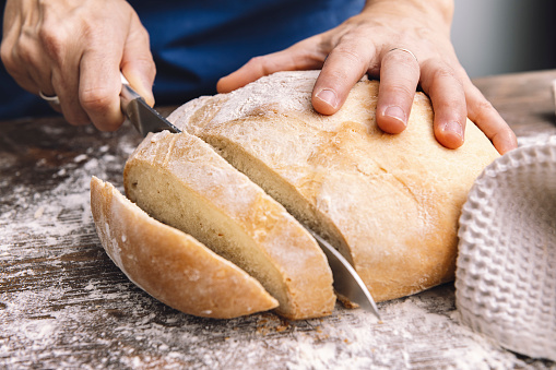 detail of hands cutting a loaf of rustic bread in slices on a wooden table full of flour, concept of healthy food at home