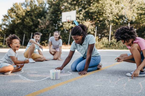 enfants avec des femmes dessinant sur le terrain de jeu utilisant la craie - animateur de colonie de vacances photos et images de collection