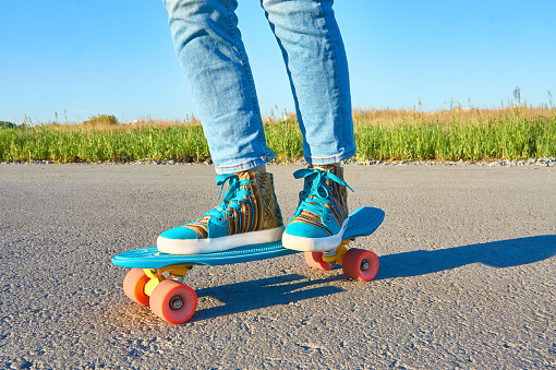 Girl in blue jeans and bright colored sneakers rides on a blue skateboard on an asphalt road on a sunny day