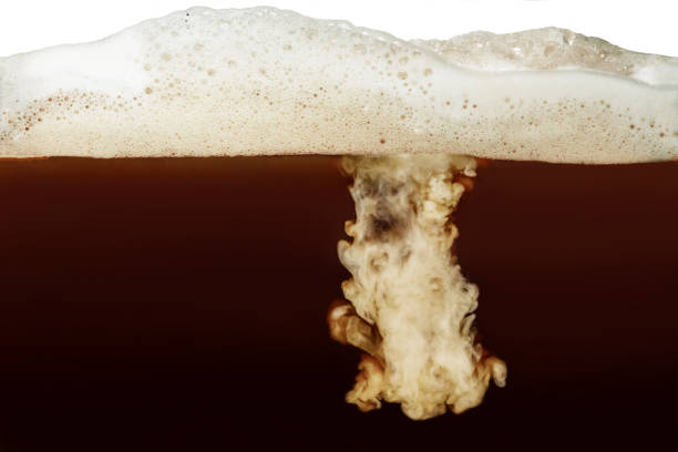 Closeup process of making latte, cappuccino, pouring milk, cream into transparent glass cup of coffee with foam. Abstract brown background with light stains. Mixing of liquids. Hot caffeine drinks.
