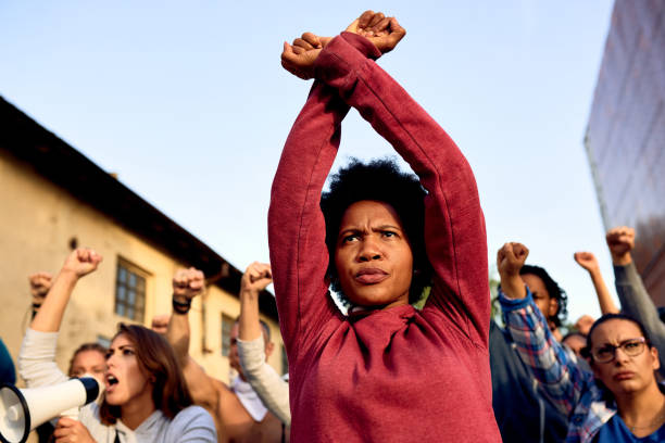Black woman with clenched fists above her head protesting with group of people on the streets. Low angle view of African American woman with arm-crossing gesture participating in a protest for human rights. police brutality photos stock pictures, royalty-free photos & images