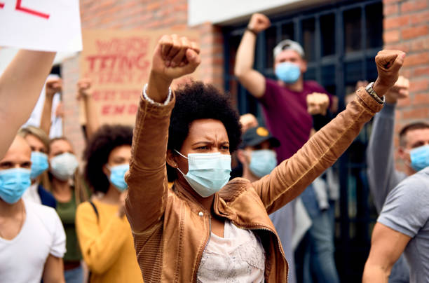 African American woman wearing protective face mask while protesting with arms raised on city streets.