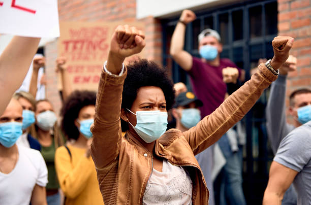African American woman wearing protective face mask while protesting with arms raised on city streets. Black woman with raised fists wearing protective face mask while supporting anti-racism demonstrations. police brutality photos stock pictures, royalty-free photos & images