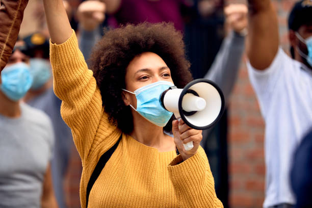 Black female activist wearing protective face mask while shouting through megaphone on a protest. Young African American woman shouting through megaphones while supporting anti-racism protests. police brutality photos stock pictures, royalty-free photos & images