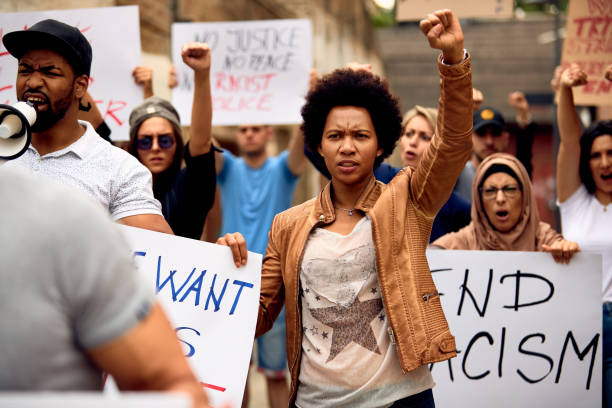 Freedom and equality for all! Multi-ethnic crowd of people protesting against racism on city streets. Focus is on African American woman with raised fist. racism photos stock pictures, royalty-free photos & images