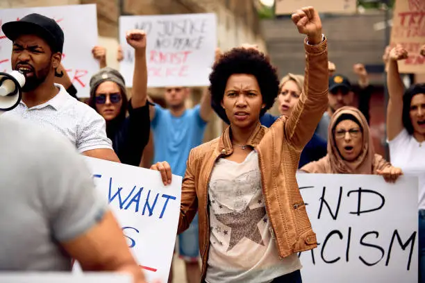 Multi-ethnic crowd of people protesting against racism on city streets. Focus is on African American woman with raised fist.