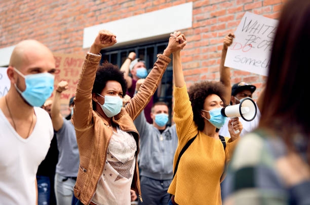 In a world of diversity, let us be united! Multi-ethnic crowd of people wearing protective face mask while protesting on anti-racism demonstrations. Focus is on two black women holding hands. police brutality photos stock pictures, royalty-free photos & images