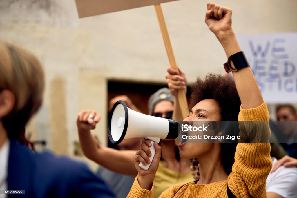 Yung black woman shouting through megaphone on anti-racism demonstrations. Young African American woman with raised fist shouting through megaphone while being on anti-racism protest. Protest Stock Photo