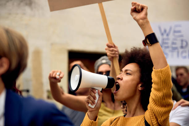 Yung black woman shouting through megaphone on anti-racism demonstrations. Young African American woman with raised fist shouting through megaphone while being on anti-racism protest. marching photos stock pictures, royalty-free photos & images
