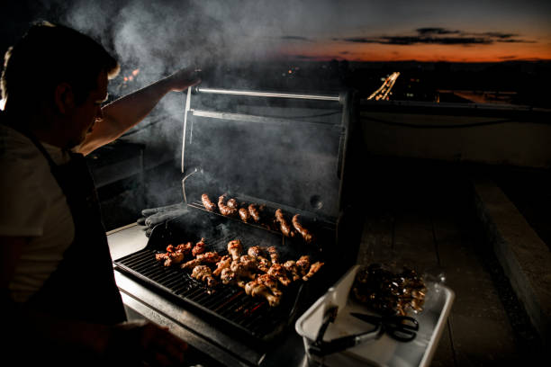 chef maschio si trova vicino al barbecue su cui vengono fritte le cosce di pollo. - spit roasted barbecue grill barbecue pork foto e immagini stock