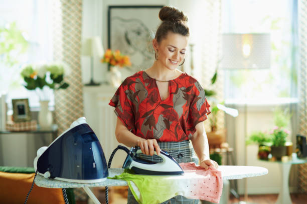 smiling housewife ironing in modern house in sunny day smiling young housewife in red blouse and grey pencil skirt ironing with steam generator on ironing board in the modern house in sunny day. stereotypical housewife stock pictures, royalty-free photos & images