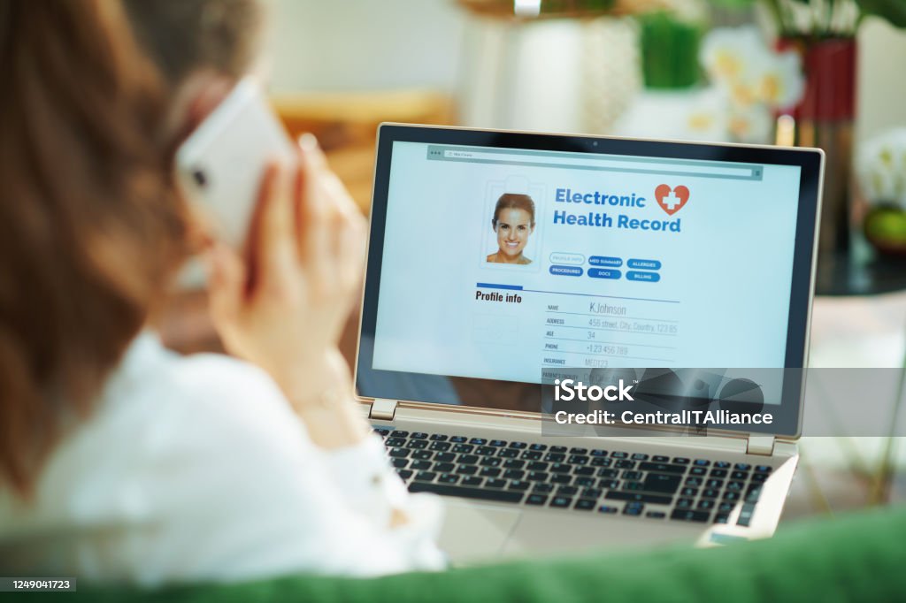 woman at home using cell phone and checking ehr Seen from behind woman at home in sunny day using a cell phone and checking electronic health record on a laptop. Electronic Medical Record Stock Photo