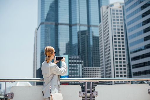 Pretty elegant businesswoman standing in the city and taking photo with her cell phone.