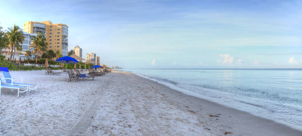 Umbrellas and chairs on Vanderbilt Beach at sunrise Umbrellas and chairs on Vanderbilt Beach at sunrise with a calm ocean and buildings along the white sand in Naples, Florida. naples beach stock pictures, royalty-free photos & images