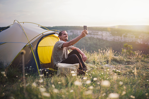 Young tourist making a selfie at sunset, he standing in front of his tent and smiling.