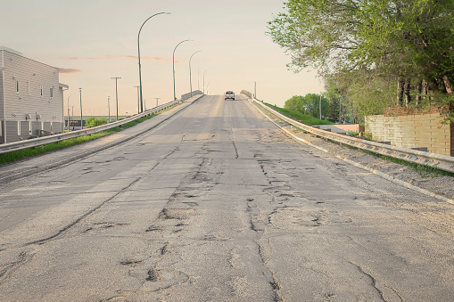 horizontal image of a paved street bridge goin uphill full of potholes and cracks and erosion under the early evening sun in the summer time.