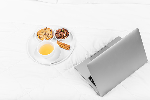 Breakfast tray on bed, with tea, muffin, cinnamon and granola, white sheets, grey laptop.