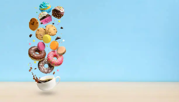Pastries, cakes and coffee cup with splash. Donuts, cookies, macaroons and coffee cup flying over blue background.