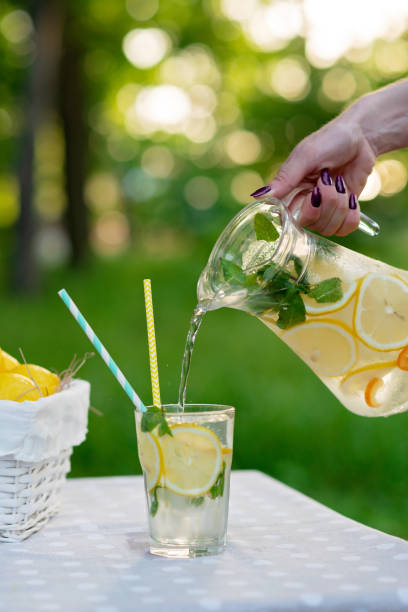 istock Female hands pour refreshing cold lemonade with lemons in ice and mint from a jug into a glass on a summer garden table on a picnic. Soft selective focus. 1249102816