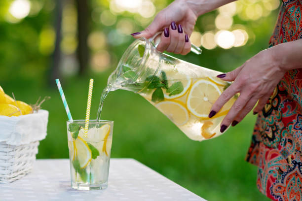 istock Female hands pour refreshing cold lemonade with lemons in ice and mint from a jug into a glass on a summer garden table on a picnic. Soft selective focus. 1249102820